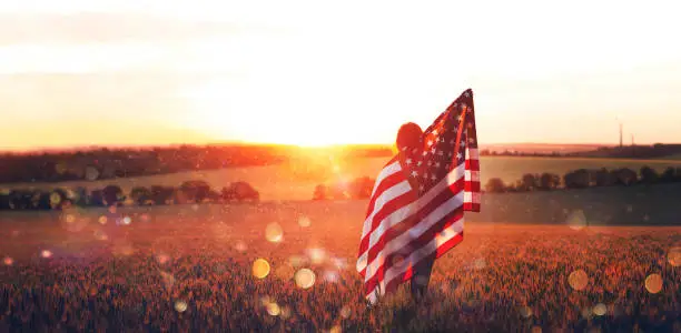 Photo of Independence Day. Beautiful young woman with the American flag in a wheat field at sunset with bokeh and sparkle. 4th of July.
