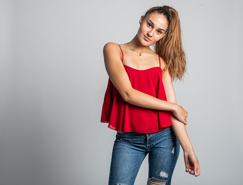 A fashionable young woman with long hair has a good time dancing and laughing in a studio photo session on a grey backdrop. She wears street style informal and colorful clothes, red pants, on some photos a red top and grey jumper.