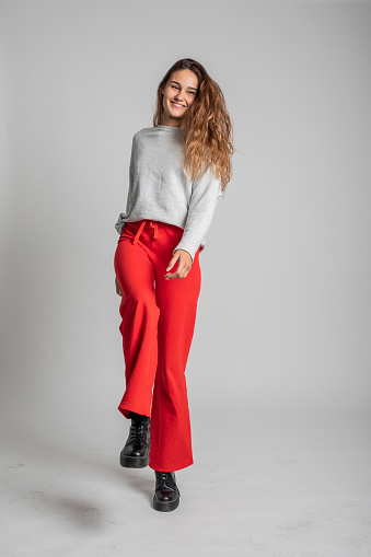 A fashionable young woman with long hair has a good time dancing and laughing in a studio photo session on a grey backdrop. She wears street style informal and colorful clothes, red pants, on some photos a red top and grey jumper.