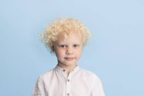 Curly and cute. Portrait of preschool boy posing isolated over blue studio background. Blond kid in casual white clothes. Concept of facial expressions, childhood, education, family. Copy space for ad