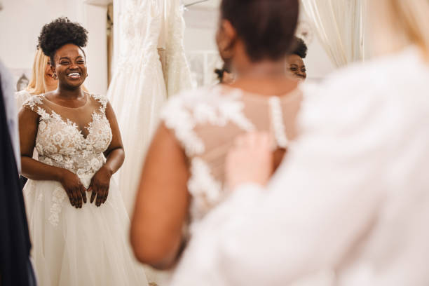 Shop assistant helping bride get into wedding dress The future bride looks around the shop and tries on wedding dresses in the presence of her best friends. Lifestyle shopping concept, post-Covid-19 era wedding dresses stock pictures, royalty-free photos & images