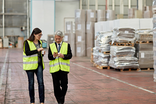Front view of mature male and female supervisors in reflective vests talking and checking smart phone as they walk together in distribution center.