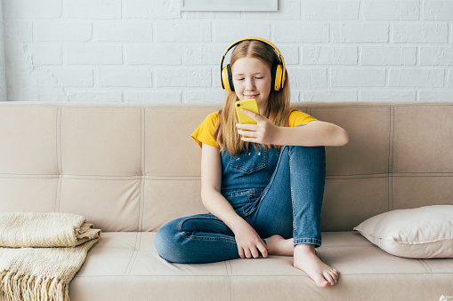 Teenage girl sitting on the couch plays the phone and listens with wireless headphones.  Summer school holidays. Time for mobile games. Video call, online communication, social networks, friends.