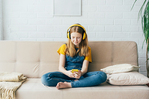 A cute teenage girl puberty sits on the couch at home and smiling looks into the mobile phone and listens to the audio in headphones. Mock up and copy space for text on the wall. Modern technology.