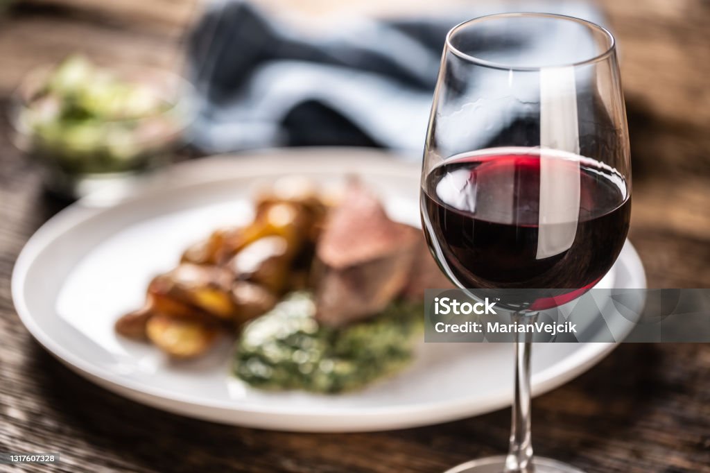 ローストポテト、ほうれん草ディップ、新鮮なサラダを添えた赤肉ステーキの典型的な平均として赤ワインを木製の机の上の白い磁器の皿に盛り付けます。 - ステーキのロイヤリティフリーストックフォト
