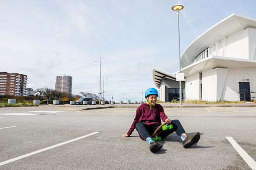 Mid-age woman skateboarding in a parking lot in Liverpool in the North of England. She is sitting down laughing on the ground after falling off her skateboard.