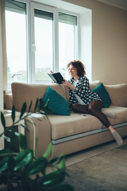 istock Charming curly haired woman is reading a book smiling on a couch at home 1317623832