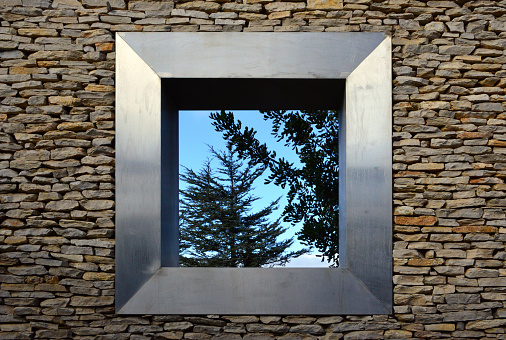 Oeiras, Portugal: looking through a square metal frame window on a stone wall - trees and sky. Wall of the Oeiras public park, Parque dos Poetas.