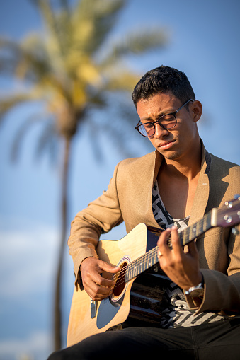 Portrait of Latin man sitting and playing guitar on street with palm trees in background