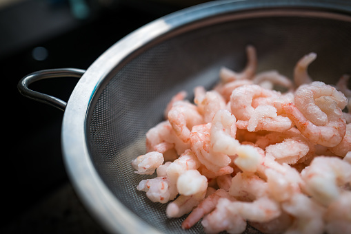 Close up image depicting a pile of frozen prawns defrosting in a metal sieve.