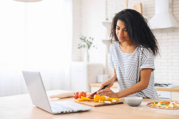 Mixed-race young woman watching cooking classes Focused mixed-race woman with afro hairstyle watching cooking classes, learning how to make yummy pizza dinner lunch watching video blog course from laptop in the modern kitchen, chopping veggies cooking class photos stock pictures, royalty-free photos & images