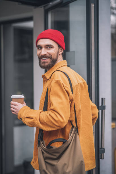 istock Man in a red hat and orange jacket with a coffee cup in hands looking cheerful 1317639984