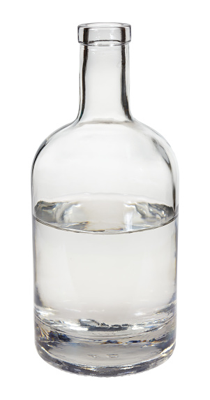 Close-up of a glass of whiskey standing on a rustic wooden table with ice cubes laying next to it. Several generic looking whiskey bottles can be seen in the background.