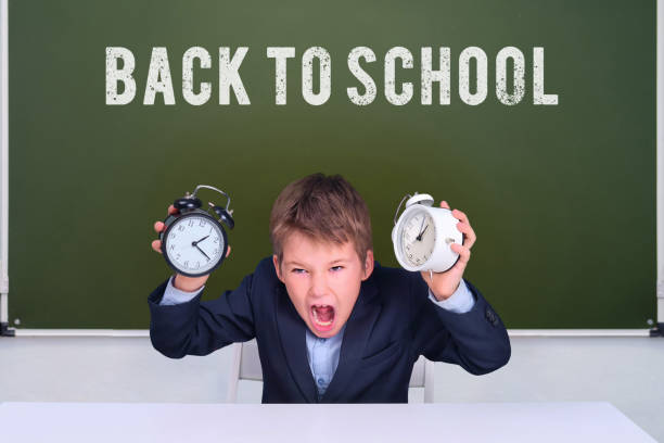 Schoolboy boy with two alarm clocks at the desk at the blackboard and the text back to school, copy space Schoolboy boy with two alarm clocks at the desk at the blackboard and the text back to school, copy space blackboard child shock screaming stock pictures, royalty-free photos & images