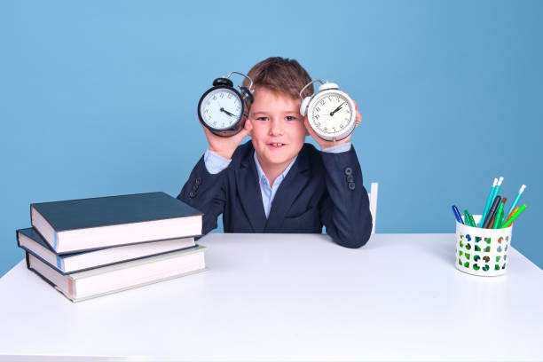 istock Happy junior boy with a clock in his hands sits at a school desk, copy space 1317650267