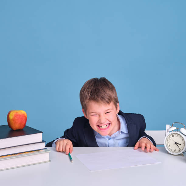 istock Schoolboy boy in uniform with textbooks and pen laughs while sitting at a school desk, blue background 1317650325