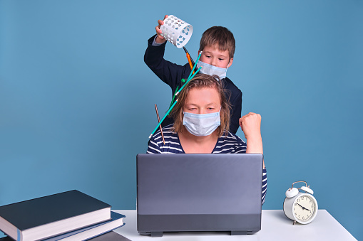 The child interferes with the mother behind the laptop at remote work in the face mask