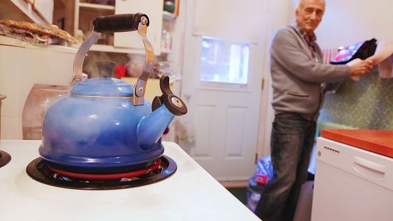 Senior man watches steaming whistling kettle from stove top prior to preparing his cup of tea