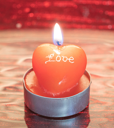 A heart shaped candle with a flame and the inscription LOVE on a blurred red background