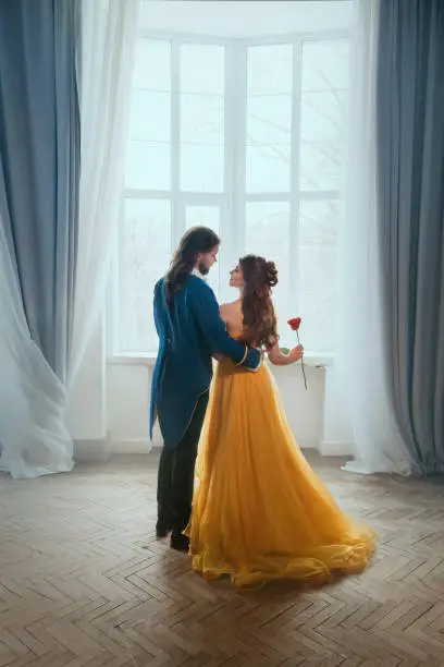Photo of Happy couple embrace in white room old classic window. Happy beauty sexy woman fantasy princess in yellow dress and man prince. Romantic male king hugs girl. Art Vintage style costume. Girl rear view