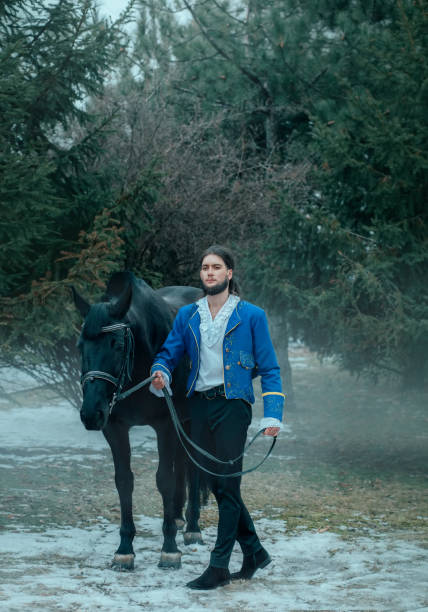 prince bel homme médiéval marche avec le cheval de course noir dans la forêt d’hiver. tient le cheval par des rênes. manteau bleu de robe bleu de costume d’époque historique, veste de caftan. guy young rider sexy king. épinette verte d’arbre - white black tree fog photos et images de collection