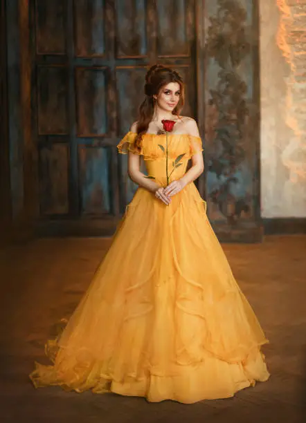 Photo of Girl beauty fantasy princess in yellow long historical, medieval silk dress holding flower red rose in her hands. Background of old gothic castle room. Fairy tale bewitched queen. Happy woman smiles