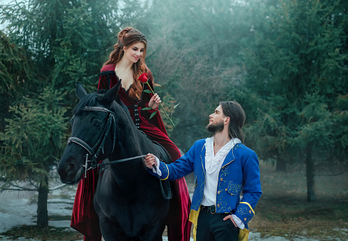 Medieval couple in love. man meets woman and gives rose in winter forest. Vintage clothing red long dress. Blue costume tailcoat caftan. Prince and princess together. Black horse. redhead hairstyle.