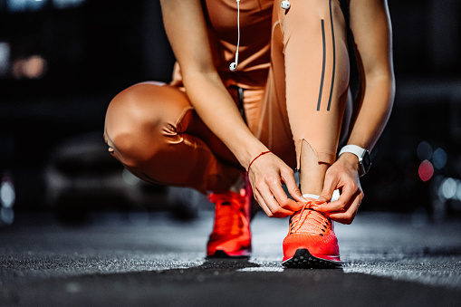 Sporty woman tying shoelace on running shoes before practice. Female athlete preparing for training in outdoors public garage. Sport active lifestyle concept. Close-up.