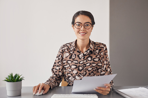 Minimal portrait of young businesswoman wearing glasses and smiling at camera while sitting at workplace holding document, copy space