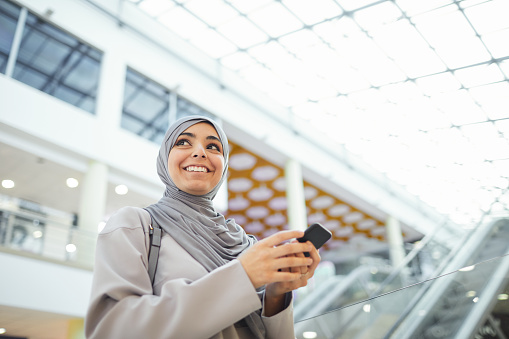 Low angle portrait of smiling Middle-Eastern woman holding smartphone while enjoying shopping in mall, copy space
