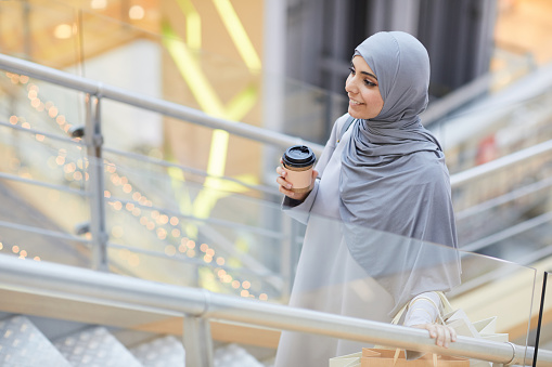 Portrait of smiling Middle-Eastern woman holding coffee cup while going up stairs in shopping mall, copy space