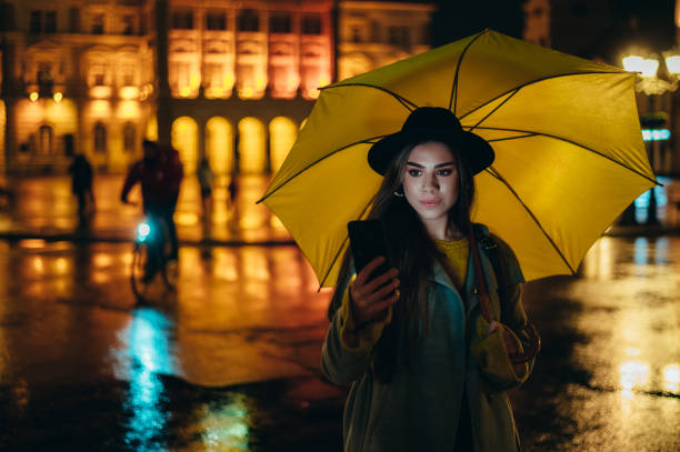 istock Young beautiful woman using a smartphone and holding a yellow umbrella during night 1317677077
