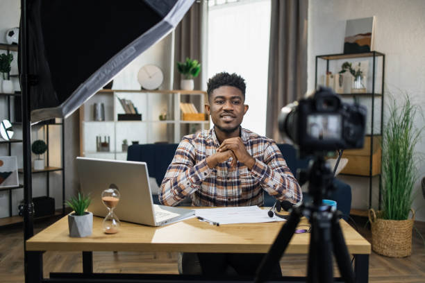 African man recording video on camera at home Handsome african man sitting at table with modern laptop and recording video on camera that fixed on tripod. Male blogger creating content for his social media. tutorial stock pictures, royalty-free photos & images