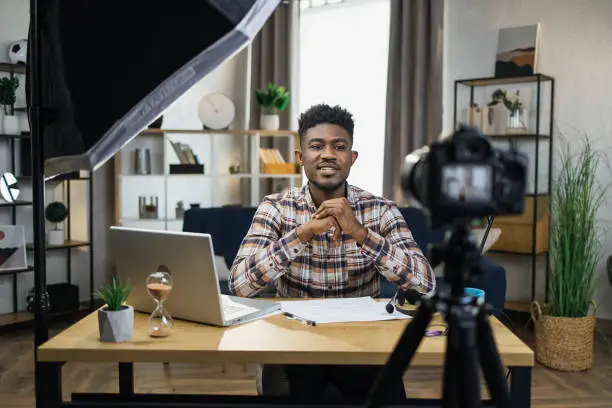 Handsome african man sitting at table with modern laptop and recording video on camera that fixed on tripod. Male blogger creating content for his social media.