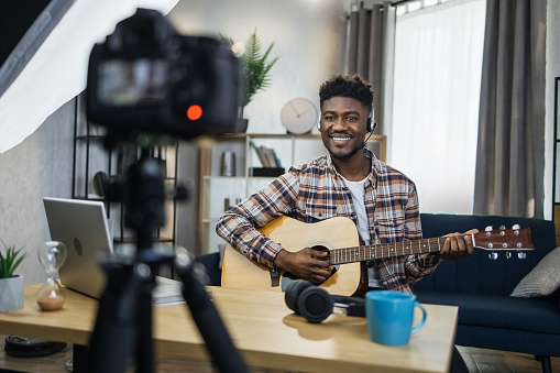 Inspired afro man playing guitar and recording music video for his social networks. Happu musician staying at home and using various modern gadgets.