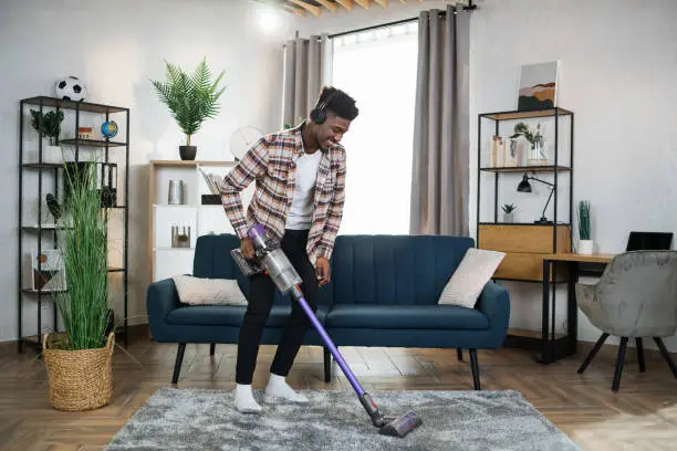Photo of Afro man in headphones cleaning carpet with vacuum cleaner
