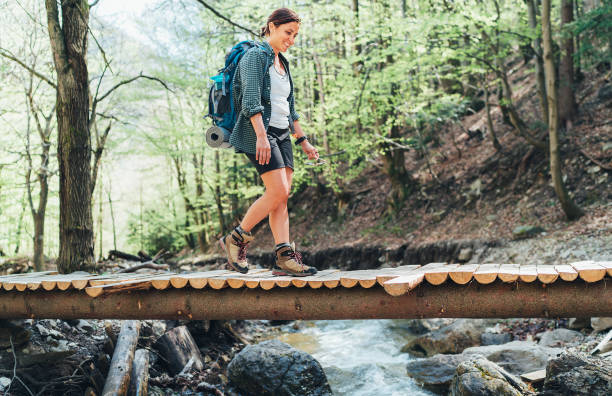 istock Smiling Middle-aged backpacker female with a backpack in trekking boots crossing mountain river bridge in the forest. Health lifestyle, traveling or trekking concept image. 1317678804