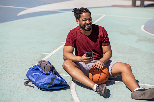 Portrait of young man sitting on the basketball field and resting from the game and using smart phone