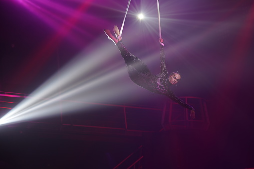 Young woman performing acrobatic element on aerial straps against dark purple background and white light