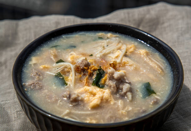 plan rapproché du congee de porc ou de la bouillie chinoise de riz avec le porc haché, l’oeuf, le champignon d’enoki et les algues dans le bol sur le napperon blanc. petit déjeuner asiatique, focuse sélectif. - thai culture food ingredient set photos et images de collection