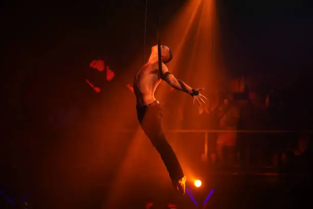 Circus artist on the aerial straps with strong muscles and naked torso balances on black background with red light