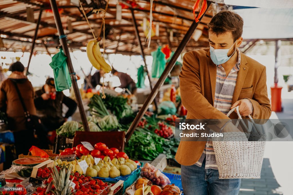 Boosting my immune system Photo of a handsome male walking through the outdoors market and caring a white wooden basket where he’s putting all fresh seasonal fruits and vegetables. He’s having a protective face mask and wearing a shirt jacket and her jeans￼￼ Immune System Stock Photo