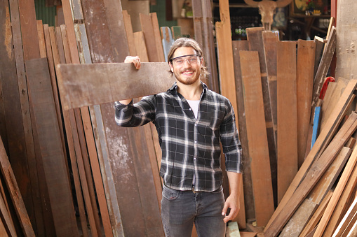 Young Caucasian carpenter man is carrying plank of wood in his own garage style workshop for hobby with copy space