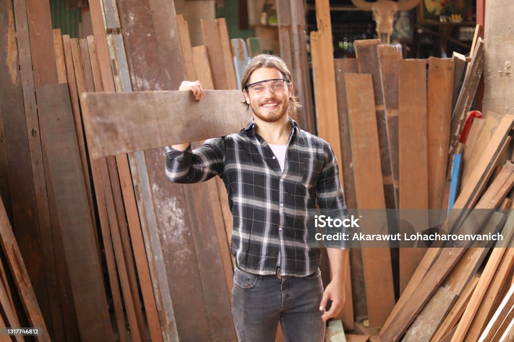 Young Caucasian carpenter man is carrying plank of wood in his own garage style workshop for hobby Young Caucasian carpenter man is carrying plank of wood in his own garage style workshop for hobby with copy space Carpenter Stock Photo