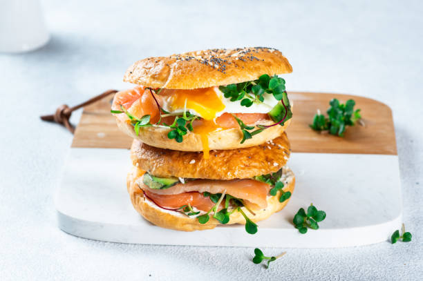 zdrowy świeżo upieczony bajgiel wypełniony łososiem, mikrozielonym, awokado i jajkiem. podawane na białym biurku. kanapka z łososiem. zdrowe śniadanie. - breakfast bread table drop zdjęcia i obrazy z banku zdjęć