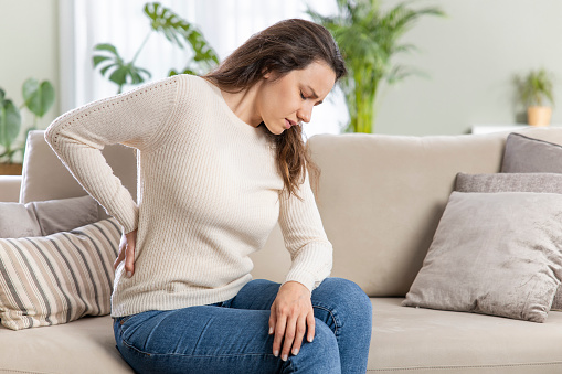 Young woman suffering with back pain, sitting on a couch and holding her lower back with hand. Axial pain, backache