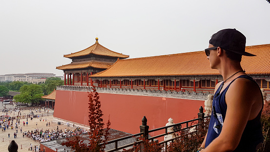 A man standing on the wall of Forbidden City in Beijing, China. There are many people in the square below. The defence wall serves also as watchtower. Discovering touristy places. Overcast