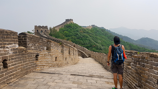 A man with hiking backpack walking around a unrenewed Gubeikou part of Great Wall of China. The wall is spreading on tops of mountains. Dense forest around it. World wonder. Tradition and history