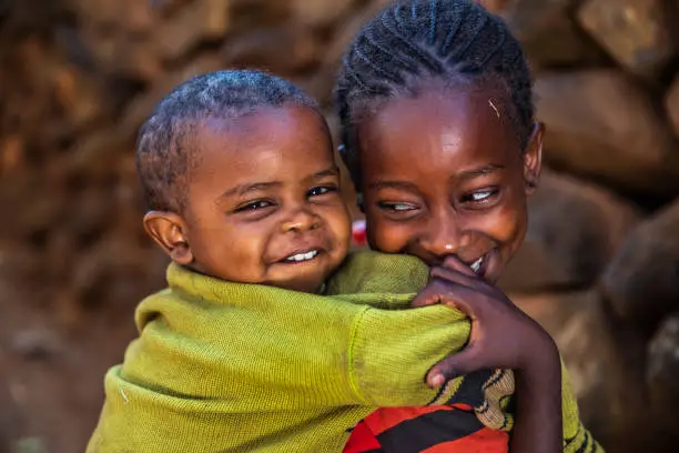 African girl carrying her younger brother. She lives in a stone village which is a part of The Konso Cultural Landscape in the Konso highlands of Ethiopia (UNESCO Heritage). Each of these villages has stone walled terraces and fortified houses.