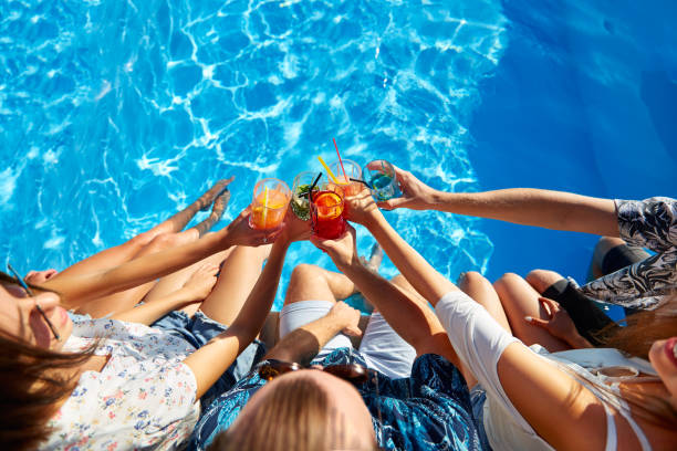 Top view of of friends clinking glasses with fresh colorful cocktails sitting by swimming pool on sunny summer day. People toast drinking beverages at luxury villa poolside party on tropical vacation stock photo
